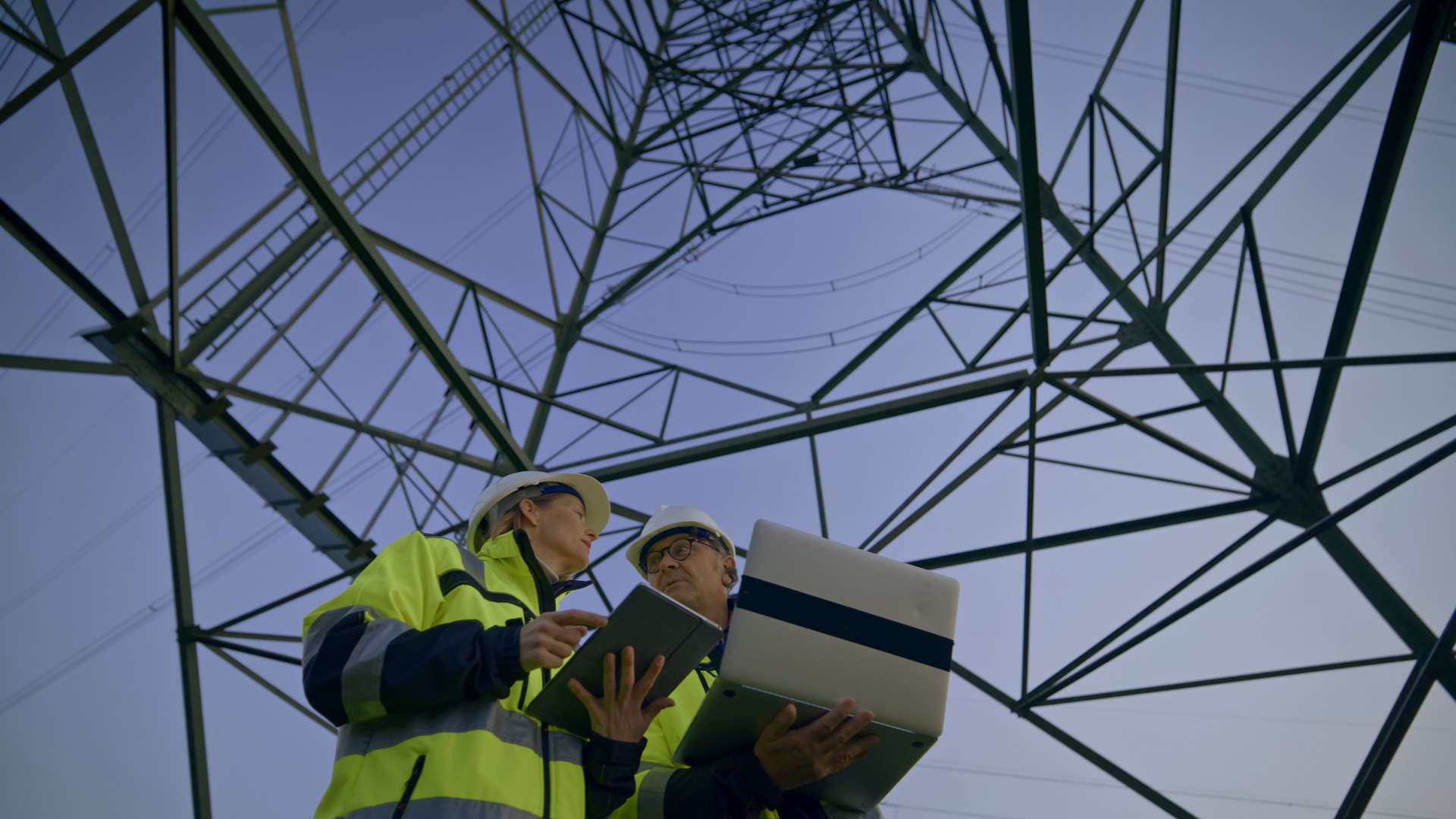 Low Angle View Of Electrical Engineers Discussing Under Electricity Pylon Against Clear Blue Sky