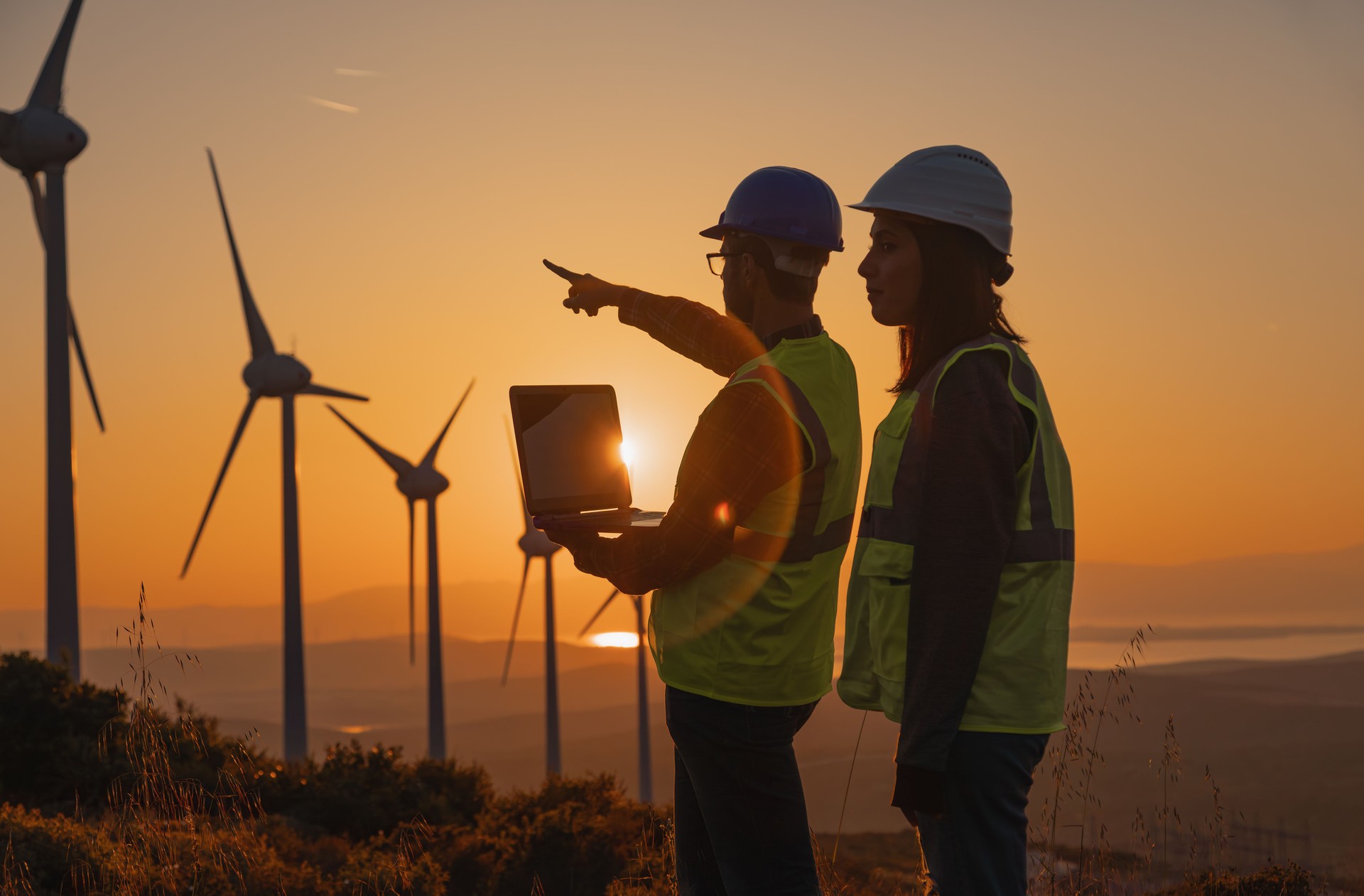 Male and Female Engineers in Safety Helmets Inspecting Wind Turbines with AI-Assisted Technology at Sunset
